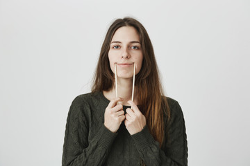 Studio shot of beautiful young woman with long straight hair in green sweater, grimacing, making herself smile with chopsticks, being displeased and upset. Face expression and negative emotions