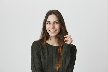 Beautiful young female with long dark hair in casual clothes smiling joyfully at camera, being pleased to hear compliments, isolated against blank wall background. Attractive european student girl