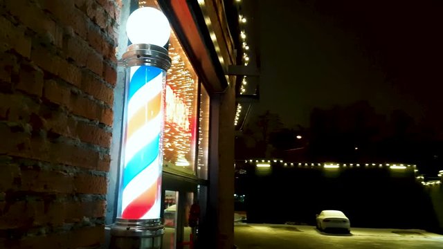 Barber's pole with a electrical motor revolving helix stripe of red white and blue colors trade sign mounted on outside wall of shop at night time