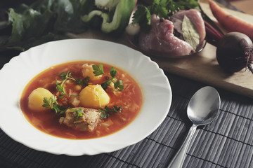 White plate with red borsch on the table near a set of fresh vegetables and meat