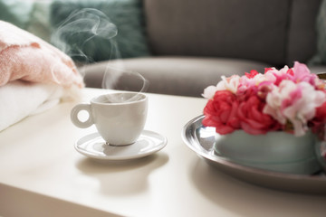 Still life details in home interior of living room. Sweaters and cup of coffe with steam on a serving tray on a coffee table. Breakfast over sofa in morning sunlight.