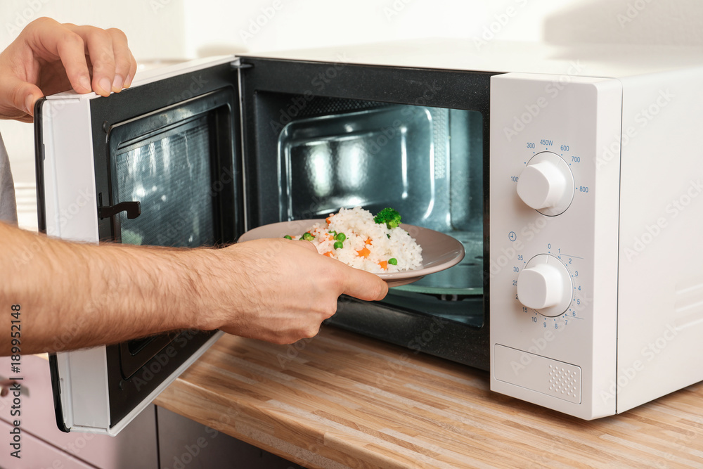 Poster man putting plate of rice with vegetables in microwave oven, closeup