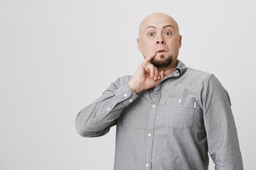 Studio shot of bald bearded man in gray shirt poses against gray background, holds little finger...