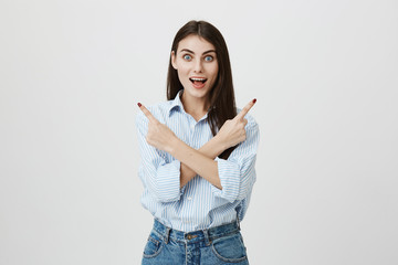 Astonished and excited young caucasian student in blue striped shirt and jeans pointing sideways with crossed hands and happy expression, standing over gray background. Woman spends great time in park