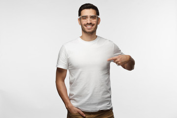 Attractive young man in transparent glasses pointing to his blank white tshirt with index finger,...