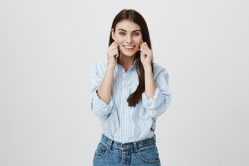 Cheerful young woman in shirt and jeans, stretching cheeks with hand and smiling broadly while looking at camera over gray background. Mother plays with her cute daughter making her laugh