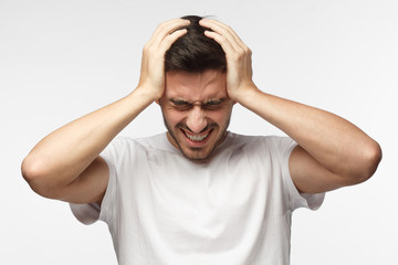 Upset unhappy young man in white t-shirt squeezing head with hands, suffering from headache. People, stress, tension and migraine concept