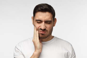 Indoor headshot of young man feeling pain, holding his cheek with hand, suffering from bad...