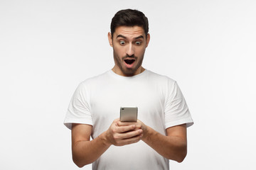 Portrait of young man isolated on gray background, looking agitated at display of her smartphone, impressed by media content from web