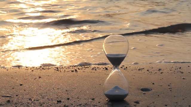 Hourglass on the beach with sea waves in bright and warm summer golden morning light showing starting time for a new day and running out in every moment