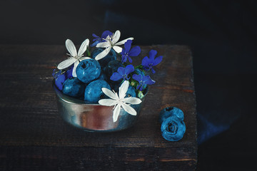 Fototapeta premium Ripe blue blueberries in a metal Cup with tiny white and blue flowers of clematis. The rustic style. Copy space.