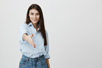 Portrait of pleasant attractive female consultant in formal shirt and jeans, stretching hand to greet customer and suggest to have a sit, standing over gray background. Nice to meet you gesture.