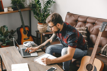 Young guitarist hipster at home with guitar browsing laptop writing