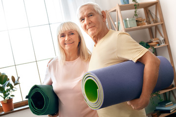 Senior couple exercise together at home health care holding yoga mats looking camera