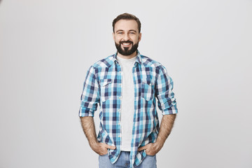 Portrait of stylish bearded guy in plaid shirt smiling broadly while holding hands in pockets, over gray background. Man at building store buy stuff to make repairment works at home