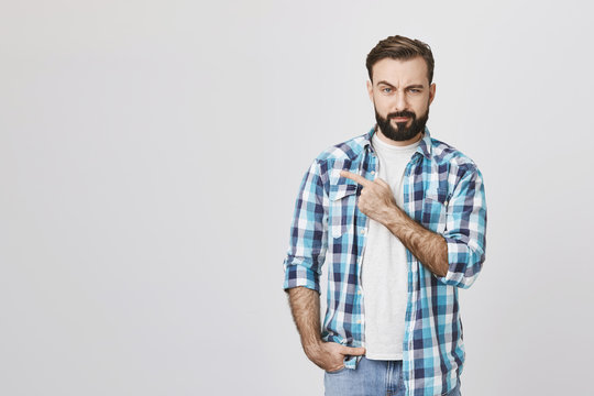 Portrait of suspicious bearded man pointing left with one hand and looking at camera, standing over gray background. Guy who went on date with girl from social network not sure it was her true photos