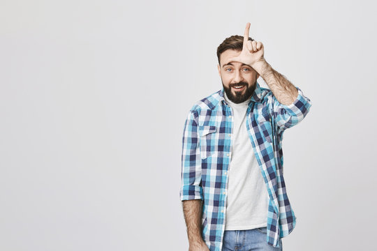 Portrait of european guy in plaid shirt holding hand on forehead showing loser gesture, smiling at camera and standing against gray background. His friend lose to him in beer pong and gloats.
