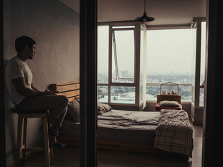 Lonely and spiritless man is sitting in his bedroom looking at sunset with smartphone on his hand.