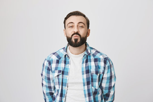 Studio shot of funny attractive adult man with beard, folding lips while waiting for kiss and being excited, standing over gray background. Guy imitates expressions his daughter makes on photos