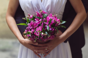 Bride and groom are holding purple rustic simple bouquet in hands together. Sunny, bright photo. White elegant dress and deep blue wedding suit.