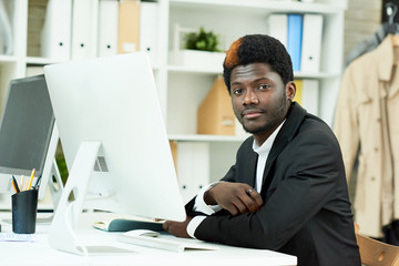 Fototapeta na wymiar Portrait of successful African-American businessman sitting at desk in modern office and smiling, looking at camera, copy space