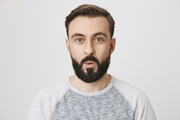 Indoor portrait of amazed bearded guy saying wow while looking at camera and standing against gray background. Man amazed by new gadget he sees in tv promotion thinking about ordering it