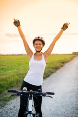 smiling girl on a bicycle with her hands up at sunset