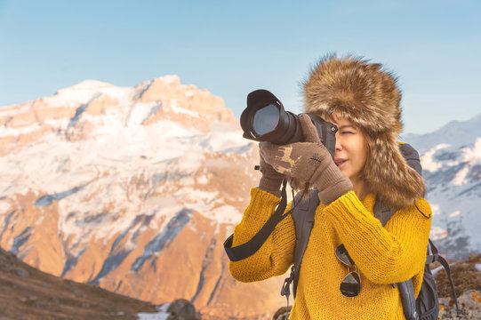 Portrait of a sweet tourist girl in a big fur hat takes pictures on her digital camera in the mountains.