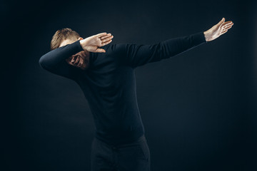 Anonymous man in black stylish outfit covering face with hands dancing on black backdrop.