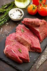 Fresh meat. Raw steak beef on a slate cutting board, tomatoes, olive oil, spices and fresh rosemary on a stone table.