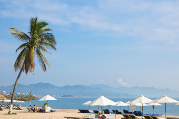 beach with palm trees, white umbrellas, beach chairs on the shore of the blue sea with mountains and city on the background