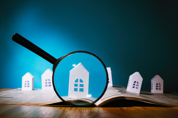 Magnifying glass in front of an open newspaper with paper houses. Concept of rent, search, purchase...