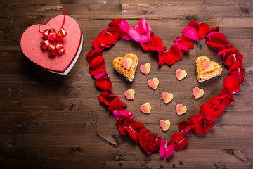 On the wooden table is a box in the form of a heart to the left of the heart of the rose petals,...