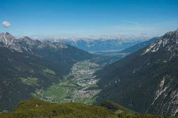 View overlooking a town in a valley between two mountain ranges. There is a small town in the valley with green space all around and forest leading up the sides of the mountains.