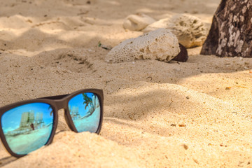 Fototapeta na wymiar Sonnenbrille im weißen Sand, in den Gläsern spiegelt sich das eine Palme, der blaue Himmel und das türkisene Meer, Steine im Hintergrund