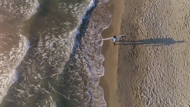 Aerial top view of young man wearing wetsuit surfer carrying surf board at the beach standing on the sand along sea shore looking at the ocean at sunset