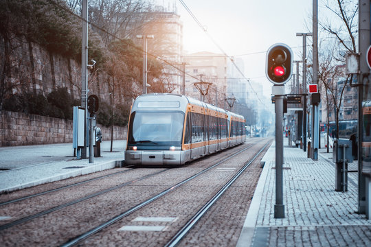 Outdoor metro or tram stop, two railway tracks, sidewalk of pavement stone, red traffic light and modern curved train with yellow stripes and huge glass windows on dull winter day, Porto, Portugal