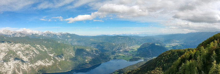 Panorama with a distant town and mount triglav in the background overlooking Lake Bohinj. The day is sunny with some clouds overhead. This is an aerial shot of the lake.