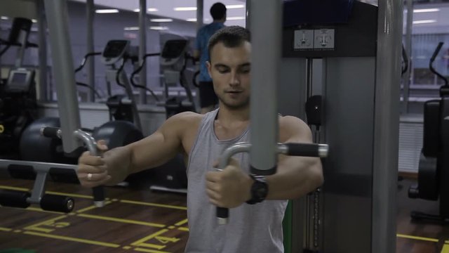 Young man is training in Pec Deck machine in gym indoors. Handsome guy spreads hands to sides, using iron equipment. Person is on intensive workout in modern sports club, performing exercise for
