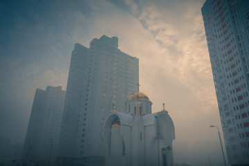 Small orthodox church at modern city residential district among high multi-storey buildings. Frosty foggy morning at city street. Kiev. Ukraine.