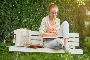 Young girl looks like a student, writes something on the white park s bench. There are some books...