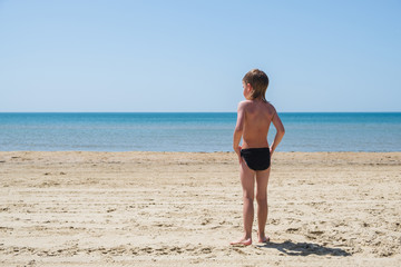 Six-year-old boy in swimming trunks stands on the beach and looks at sea.