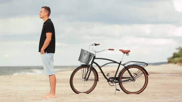 Young pretty man standing on the beach near retro bicycle. Handsome guyl resting after cycling near the sea.