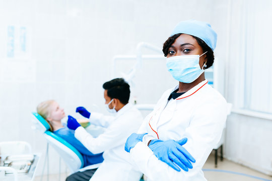 Young African american female dentist in mask in front of dentist checking teeth to patient at clinic. Dental clinic concept.