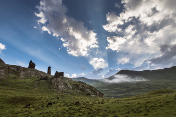 Fototapeta na wymiar Summer morning at Caucasus, Tusheti region , Georgia. Hills covered with green grass. On the high rocks located ruins of medival tushetian defensive tower. Sunrays highlighting clouds in the blue sky