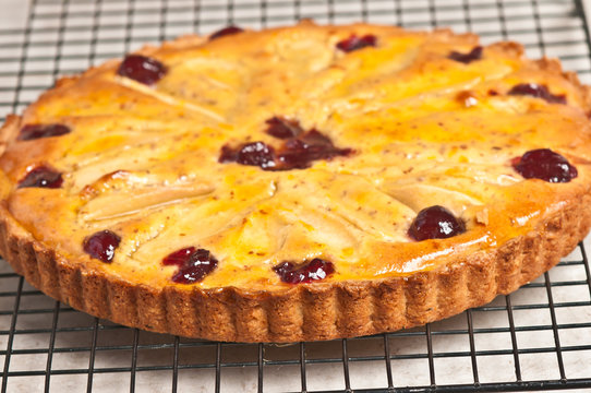 Close up, top view of a freshly baked, homemade, pear cranberry tart with almond glaze cooling on a wire  cooling rack on a wood serving board with a glass bowl of glaze and a wood application brush