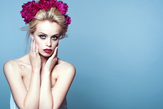 Portrait Of Beautiful Blond Model With Updo Hair Blown By The Wind And Perfect Make Up Wearing Peony Head Garland Holding Her Hands At Her Face. Isolated On Blue Background. Copy Space