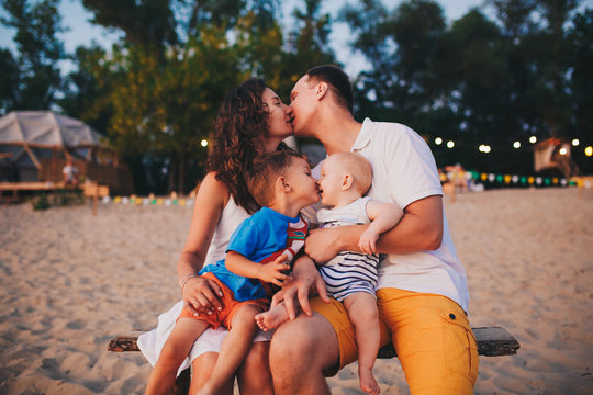The concept of a family vacation. Young family and two sons sitting on a bench in the evening on a sandy beach. Mom and Dad kiss, the older brother kisses the younger on the lips