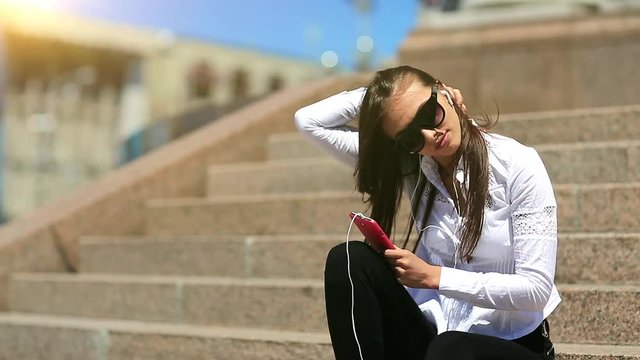 Young woman sits on the stairs and listens to music. Beautiful girl with long hair in sunglasses with red smartphone listens to music in the city centre