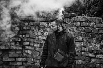 Vape man. Photo of a handsome young white guy with modern haircut in the black clothes vaping and letting off steam from an electronic cigarette near old destroyed brick wall. Black and white photo.